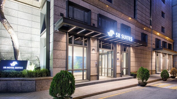 Exterior del SR Hotel Suites con una entrada moderna, amplias puertas de vidrio y plantas en macetas.