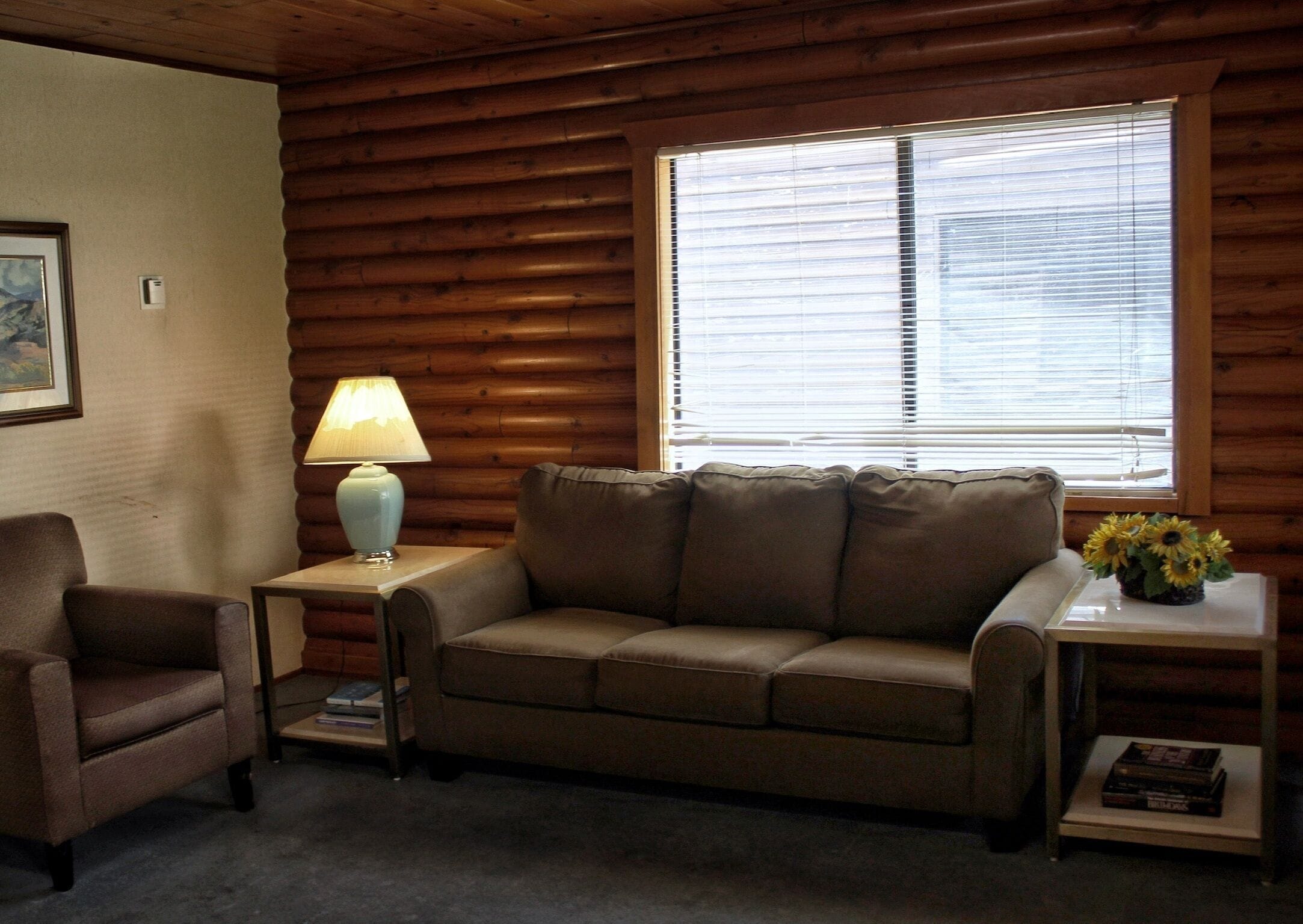 L'intérieur d'une cabane en rondins, avec un canapé en cuir marron, une petite table avec une lampe et une fenêtre équipée de stores.