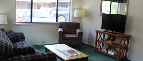 A living room with a sofa, armchair, coffee table, TV stand, and a window with a view of parked cars.
