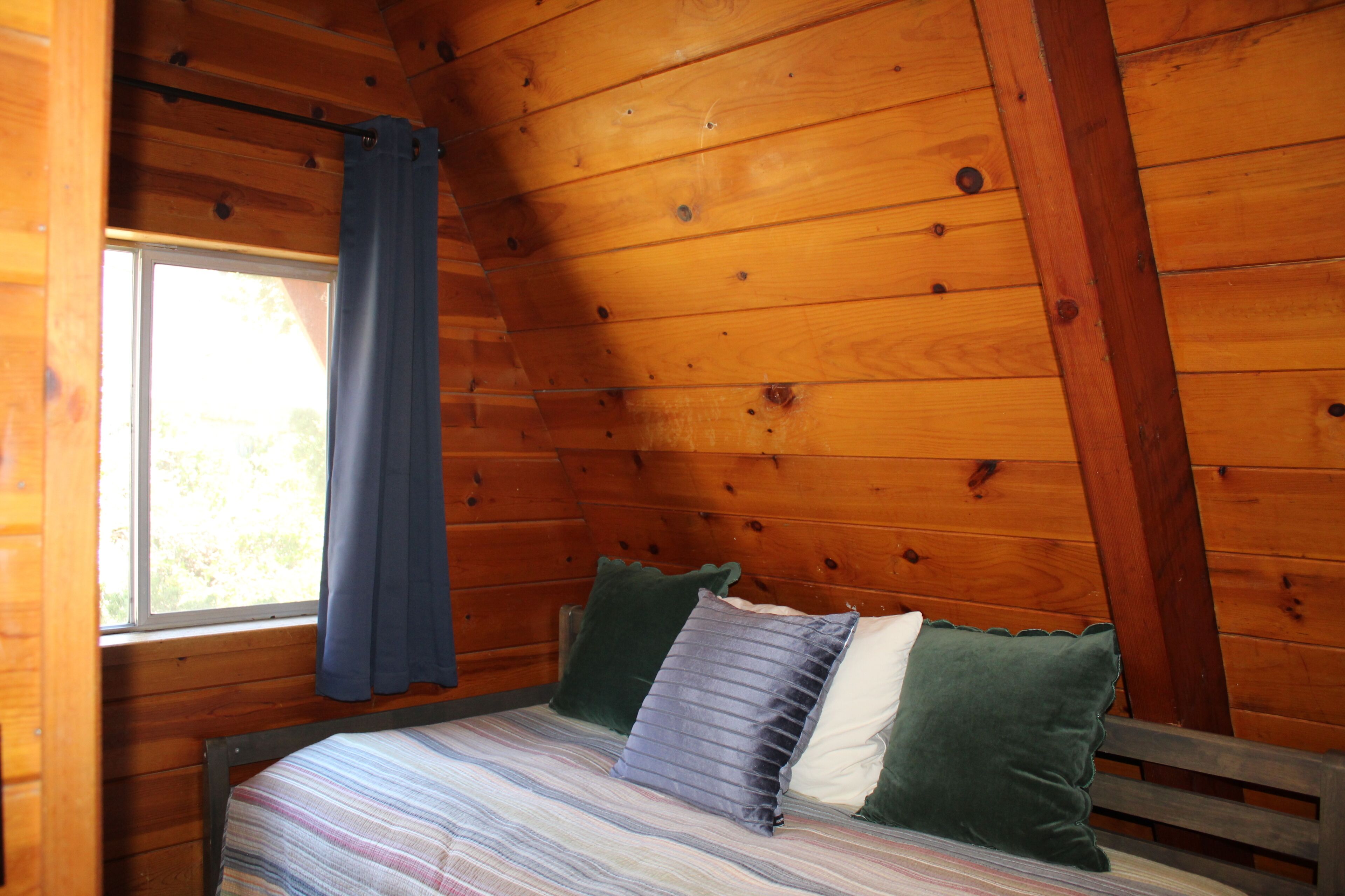 A wooden cabin room with a bed, a window with curtains, and a sloped ceiling.