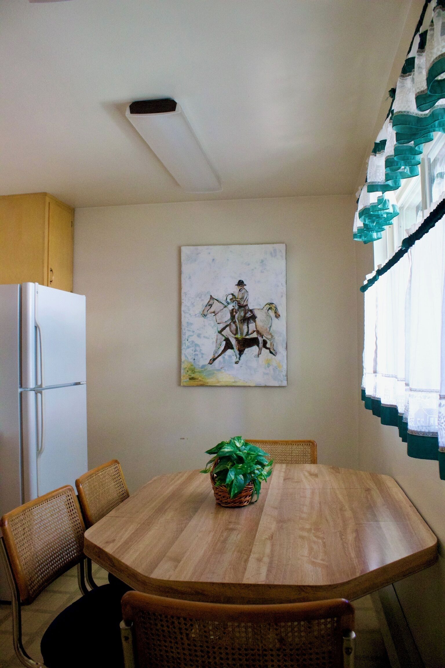 Un coin repas avec une table en bois, des chaises en osier, un réfrigérateur et un tableau accroché au mur.