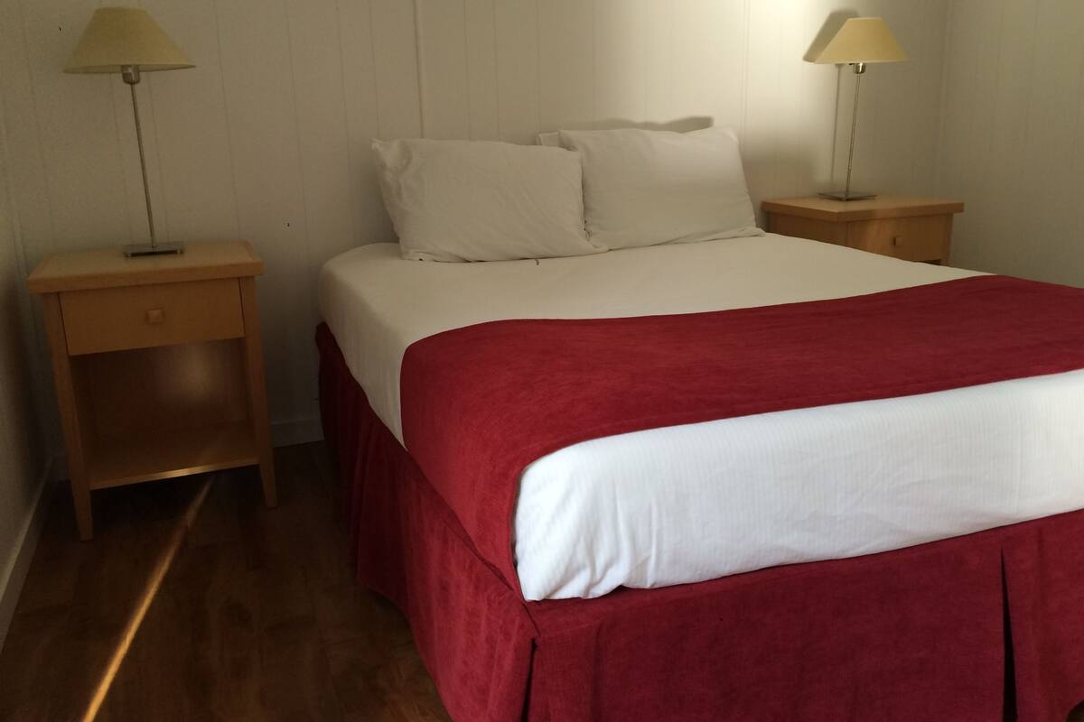 A neatly made bed with a red bedspread and white pillows, flanked by two bedside tables with lamps.