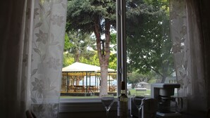 A table with a wine glass, a bottle, and a coffee maker, with a view of an outdoor seating area through a window with floral curtains. - Kokanee Beach Resort Motel (Lake Country)