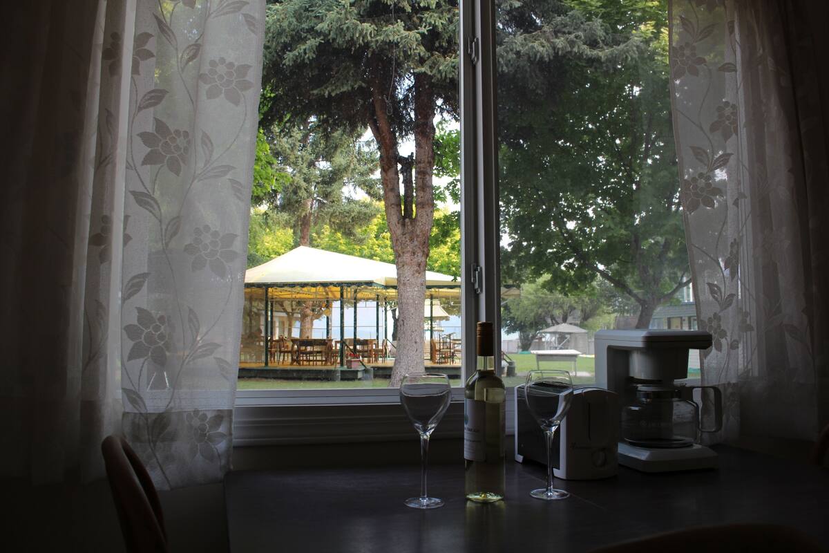 A table with a wine glass, a bottle, and a coffee maker, with a view of an outdoor seating area through a window with floral curtains.
