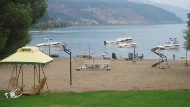 A lakeside beach with boats, a swing chair, and a table with chairs. - Kokanee Beach Resort Motel (Lake Country)