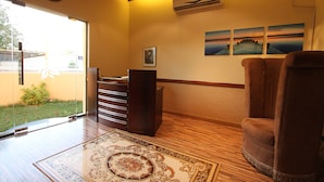 A room with a wooden floor, a brown armchair, a desk with drawers, a clock on the wall, and a window with a view of greenery. - Al Maha Residence RAK (Ras Al Khaimah)