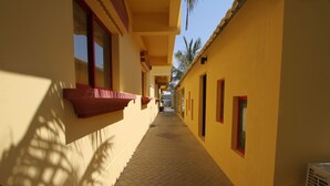 A narrow corridor with yellow walls and red-framed windows, leading to a covered area with palm trees. - Al Maha Residence RAK (Ras Al Khaimah)