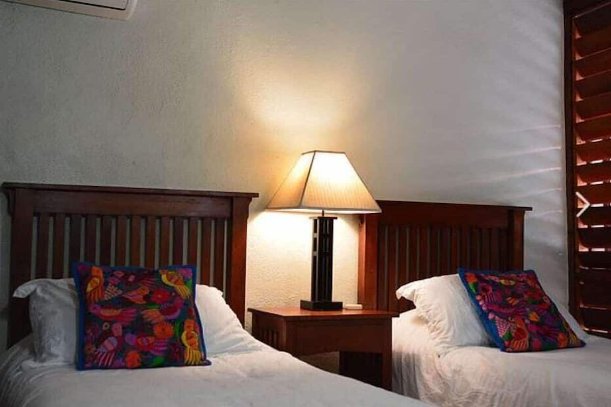 Dos camas con almohadones de colores, una mesita de noche con una lámpara y una ventana con persianas.