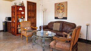 A living room with a brown leather sofa, two armchairs, a glass coffee table, a wooden bookshelf, and a painting on the wall.