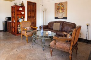 Ein Wohnzimmer mit einem braunen Ledersofa, zwei Sesseln, einem Glastisch, einem Bücherregal aus Holz und einem Bild an der Wand.