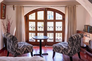 Een kamer met een groot boogvenster, twee gestreepte fauteuils, een klein rond tafeltje en een houten kast vol boeken.