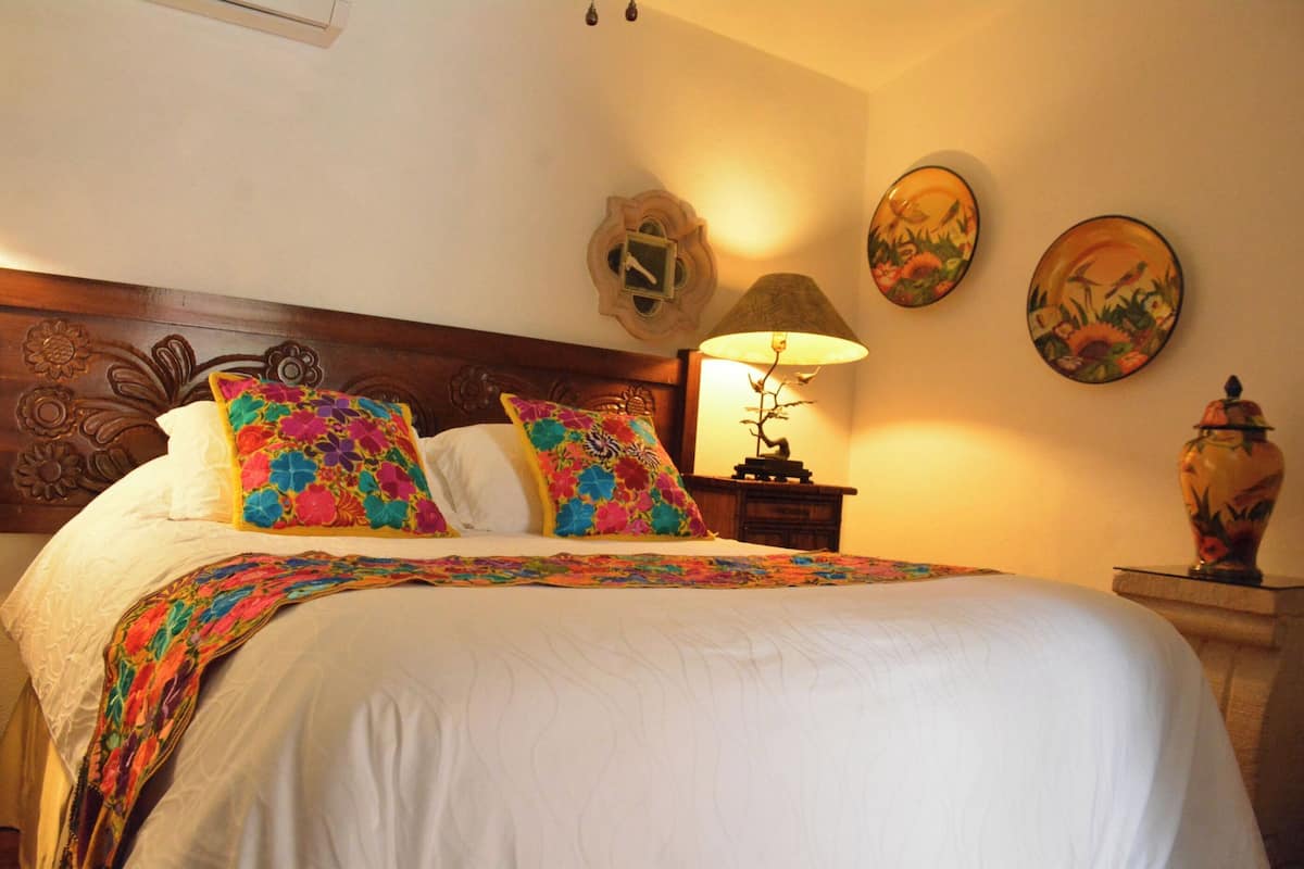 Una cama bien hecha con colcha floral y almohadas coloridas, un cabecero de madera, una lámpara de noche y platos decorativos en la pared.