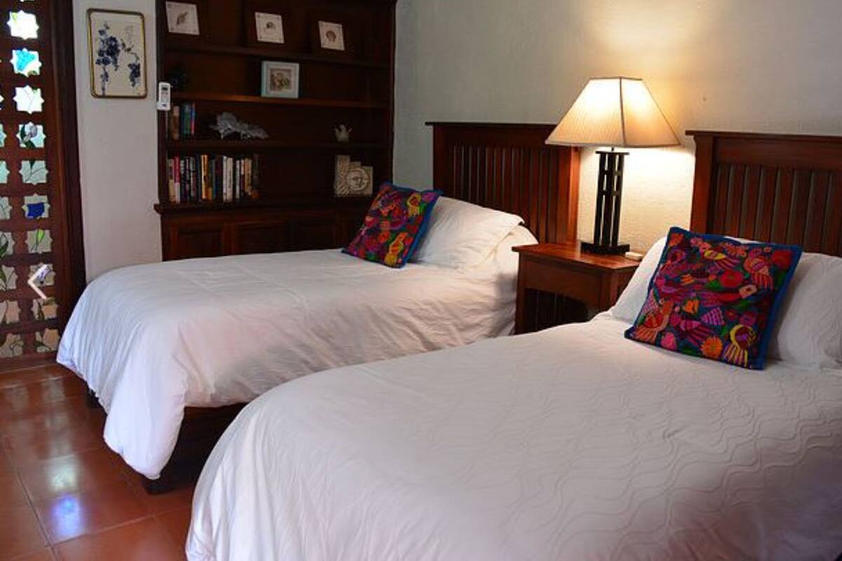 Una habitación con dos camas, una mesita de noche con lámpara, una estantería y una pared decorada con cuadros.