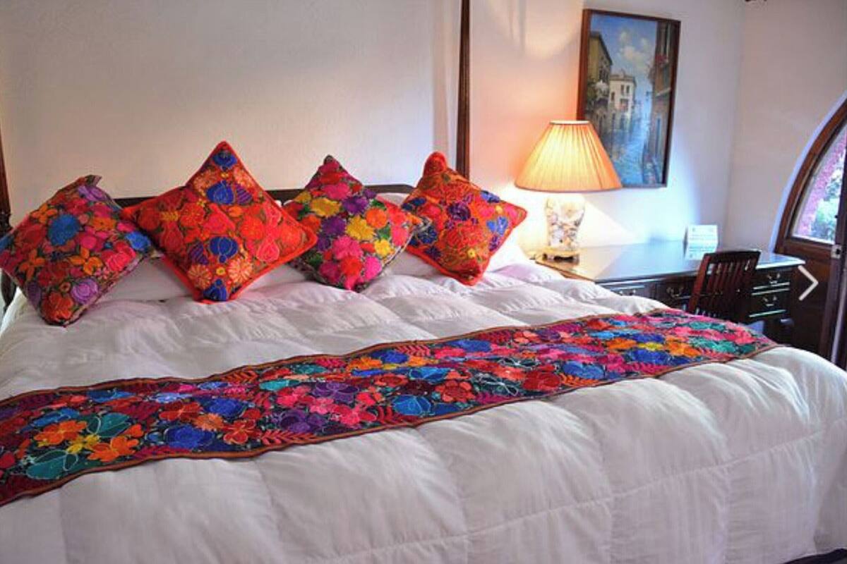 Una cama con edredón blanco y almohadas florales de colores, una lámpara y un cuadro en la pared.