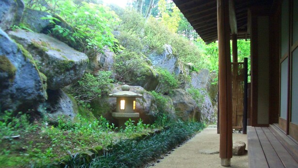 Een traditionele Japanse tuin met een stenen lantaarn, een houten pad en een gebouw met schuifdeuren.