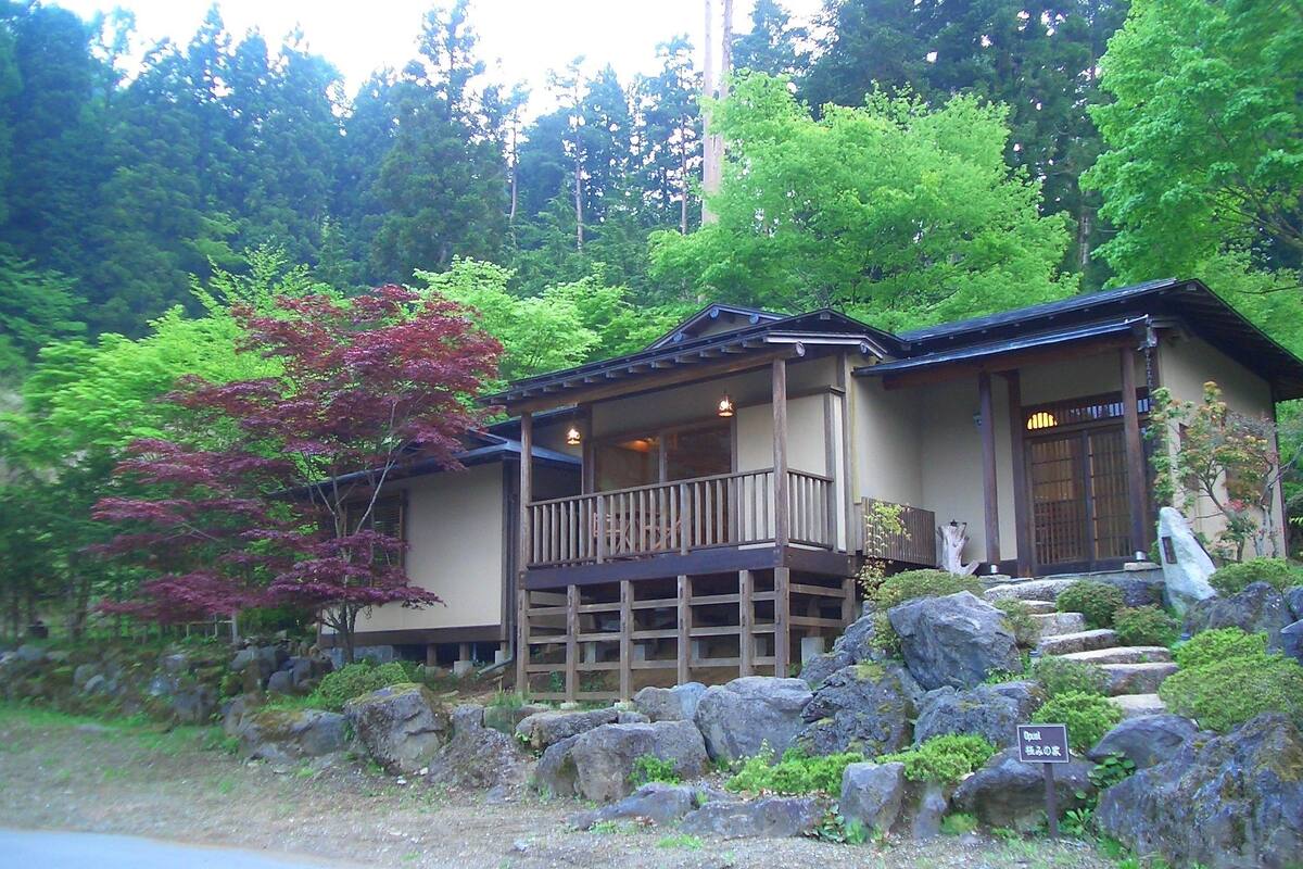 Une maison traditionnelle de style japonais, dotée d’un balcon en bois et entourée d’une végétation luxuriante et de rochers.