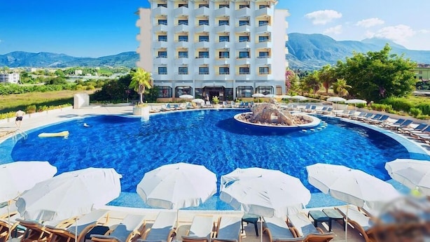 A hotel with a large swimming pool surrounded by lounge chairs and umbrellas, set against a mountainous backdrop. - Sunshine Hotel (Alanya)