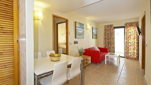 Un coin repas avec une table, des chaises et un panier de fruits. On y trouve un canapé rouge et une petite table blanche avec une lampe. La pièce est dotée d’un sol carrelé et d’une porte en bois.