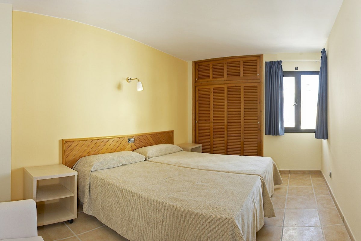 Une chambre avec deux lits, une table de chevet, une lampe fixée au mur, une armoire en bois et une fenêtre avec des rideaux.