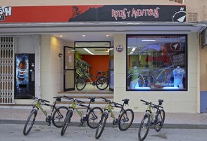 Una fachada con un cartel que dice "Rutas y Aventura" y un amplio escaparate que muestra bicicletas y una camiseta de ciclismo.