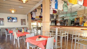 Un restaurant dont les tables sont recouvertes de nappes rouges, avec des chaises blanches et un coin bar doté d’un comptoir.