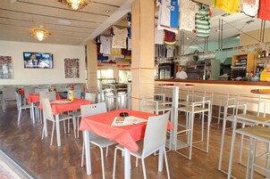 Un restaurante con mesas cubiertas de manteles rojos, sillas blancas y una zona de bar con mostrador.