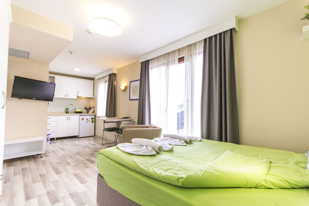 A hotel room with a green bed, a TV, a desk, and a kitchenette.