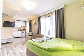 A hotel room with a green bed, a TV, a desk, and a kitchenette.