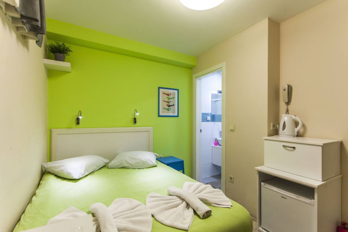 A hotel room with a green bedspread, white pillows, a small potted plant, a framed picture, a white telephone, and a white cabinet with a kettle.
