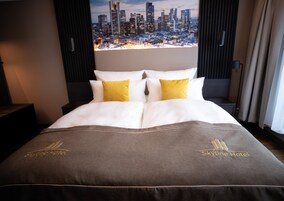 Habitación de hotel con una cama que ofrece vistas panorámicas de la ciudad a través de la ventana, un cabecero con líneas verticales y una colcha con el nombre del hotel bordado.