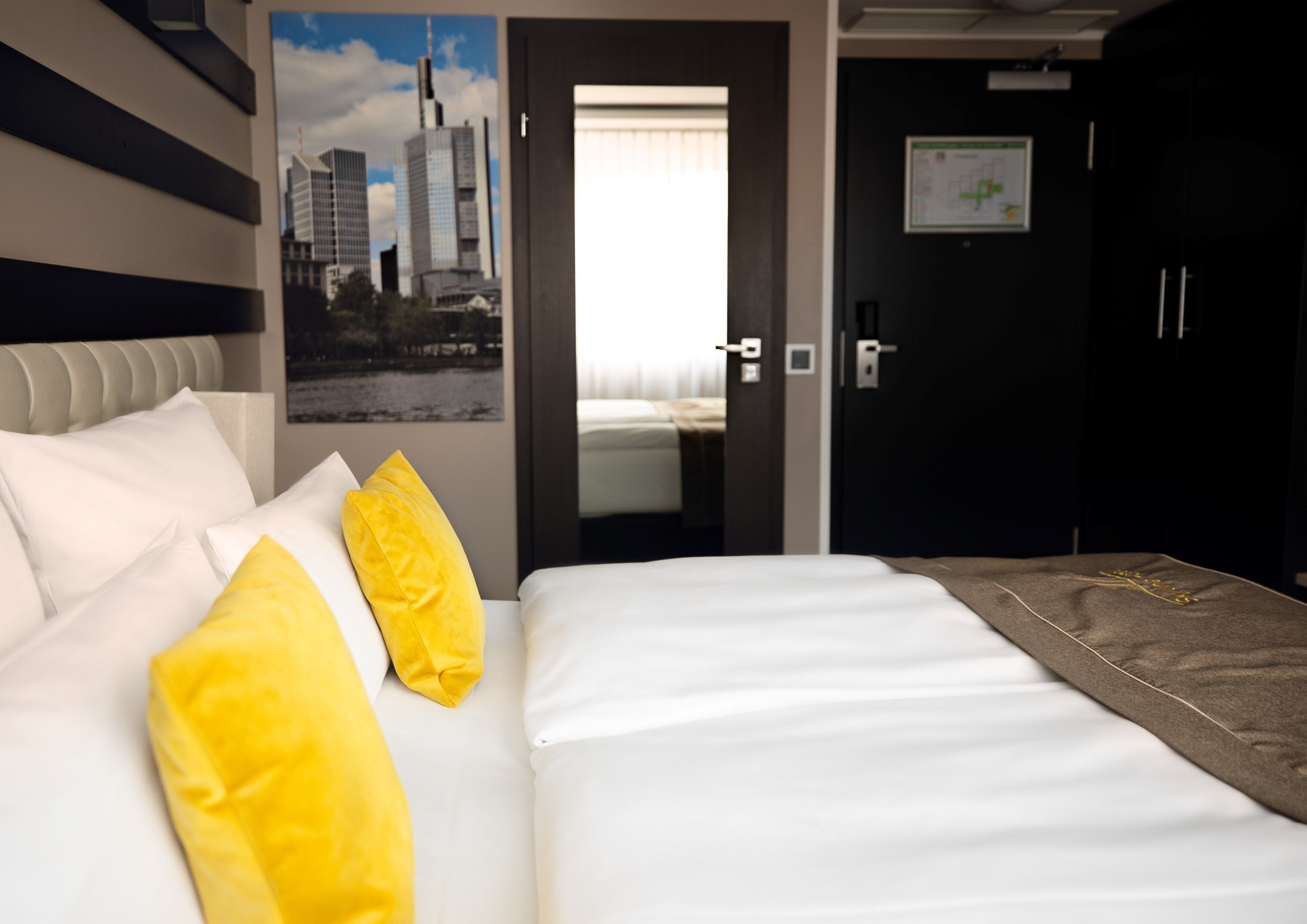 A hotel room with a bed, white and yellow pillows, a view of a cityscape through an open door, a dark wardrobe, and a framed picture on the wall.