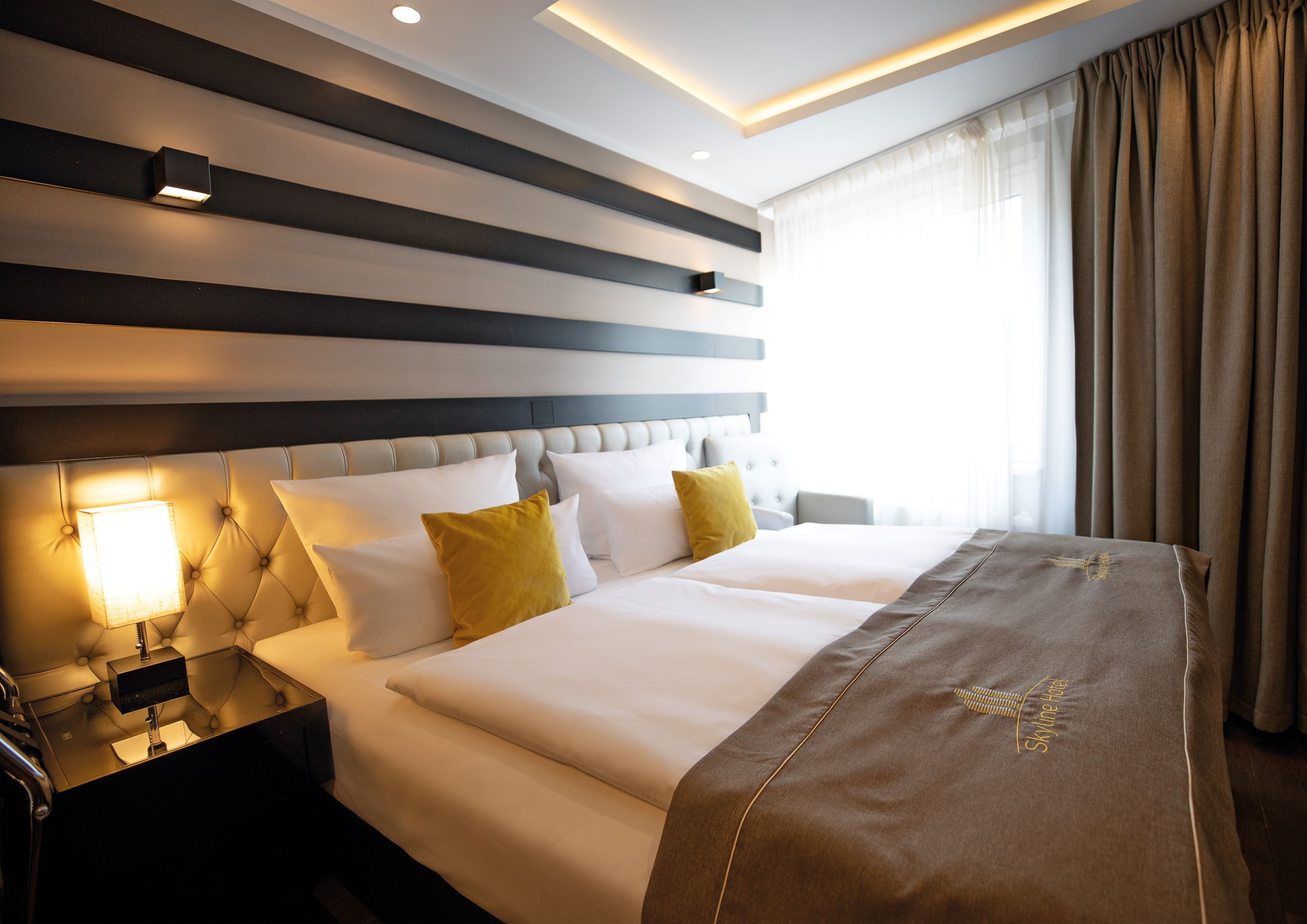 Ein Hotelzimmer mit einem großen Bett, einem Nachttisch, einer Wandleuchte und einem Fenster mit Vorhängen.