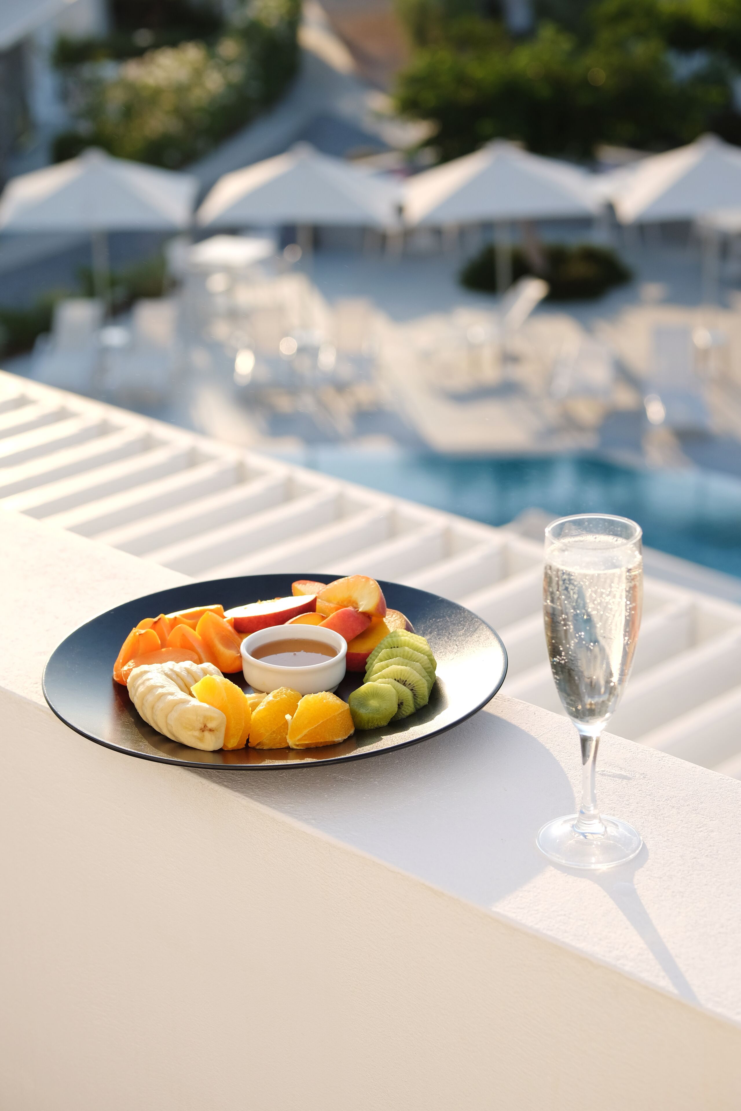 A dining table with a plate of fruit and a glass of champagne, overlooking a pool area with umbrellas.