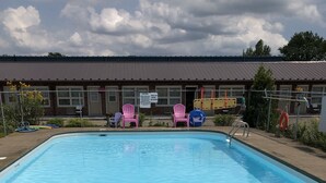 A swimming pool with clear blue water, surrounded by a fenced area with pink chairs and a building in the background. - Good Night Inn (Port Colborne)