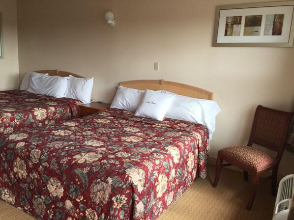 A double bed with floral bedding, a wooden headboard, and white pillows. There is a wooden nightstand with a lamp, a chair, and a framed picture on the wall. - Good Night Inn (Port Colborne)
