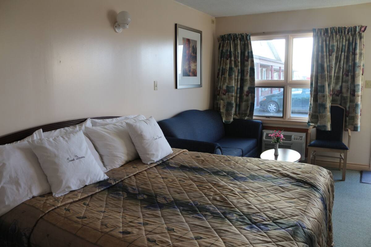 Une chambre d’hôtel avec un grand lit, un fauteuil bleu, une petite table avec un vase et une fenêtre avec des rideaux.
