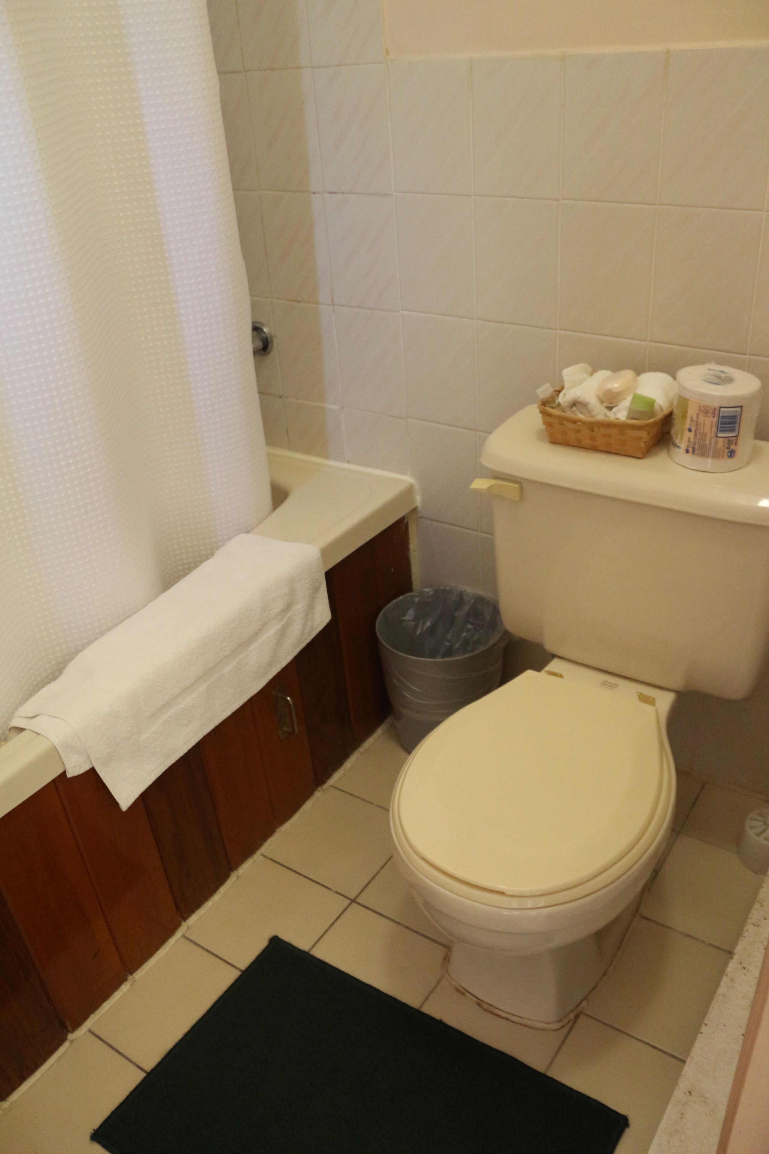 Un baño con inodoro, un bote de basura, lavamanos con toalla y un cesto de artículos de aseo sobre el tanque del inodoro.