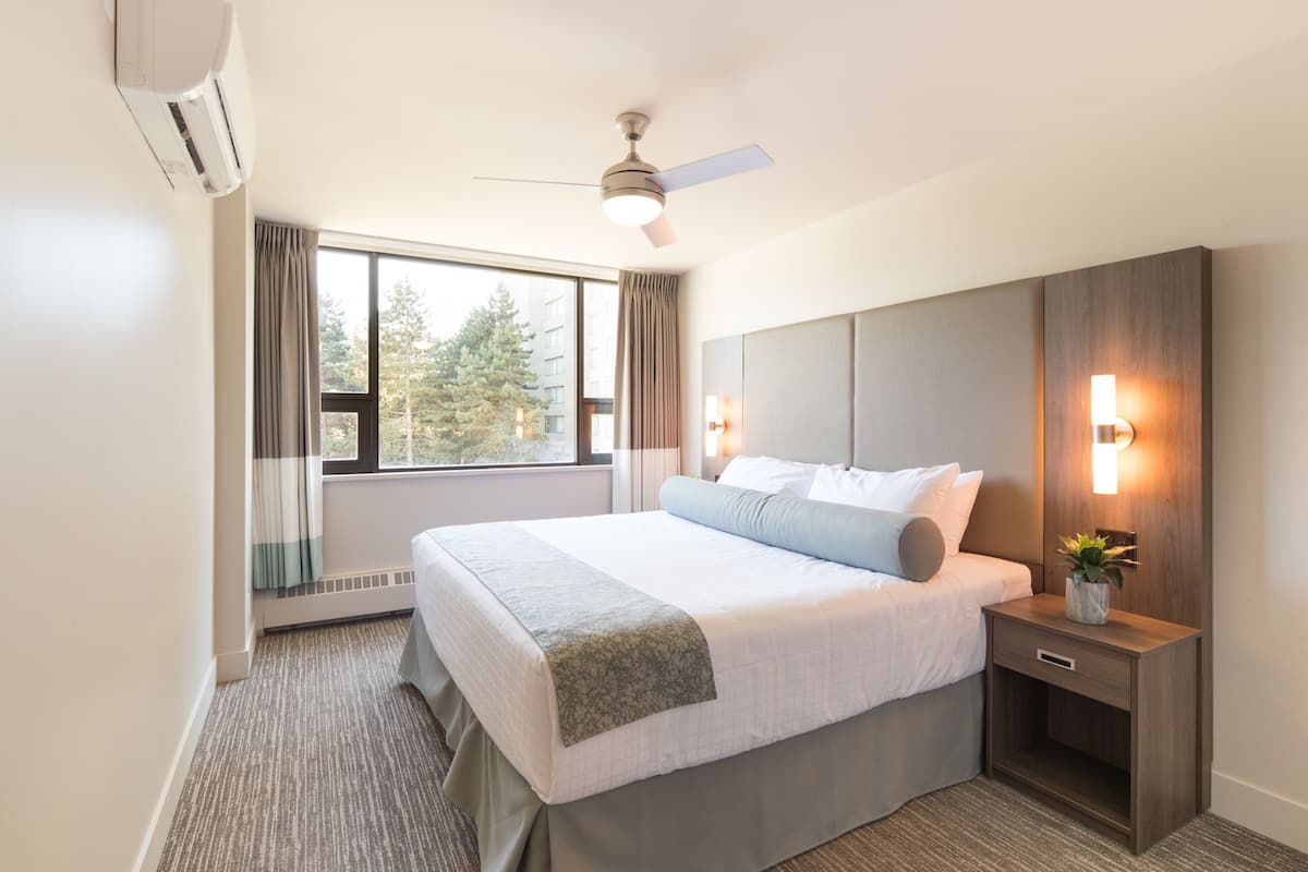 A modern hotel room with a large bed, a ceiling fan, a nightstand with a lamp, and a window with curtains.