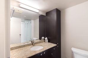 A bathroom with a granite countertop, a sink, a mirror, a dark cabinet, and a toilet. - Gage Suites at UBC (Vancouver)