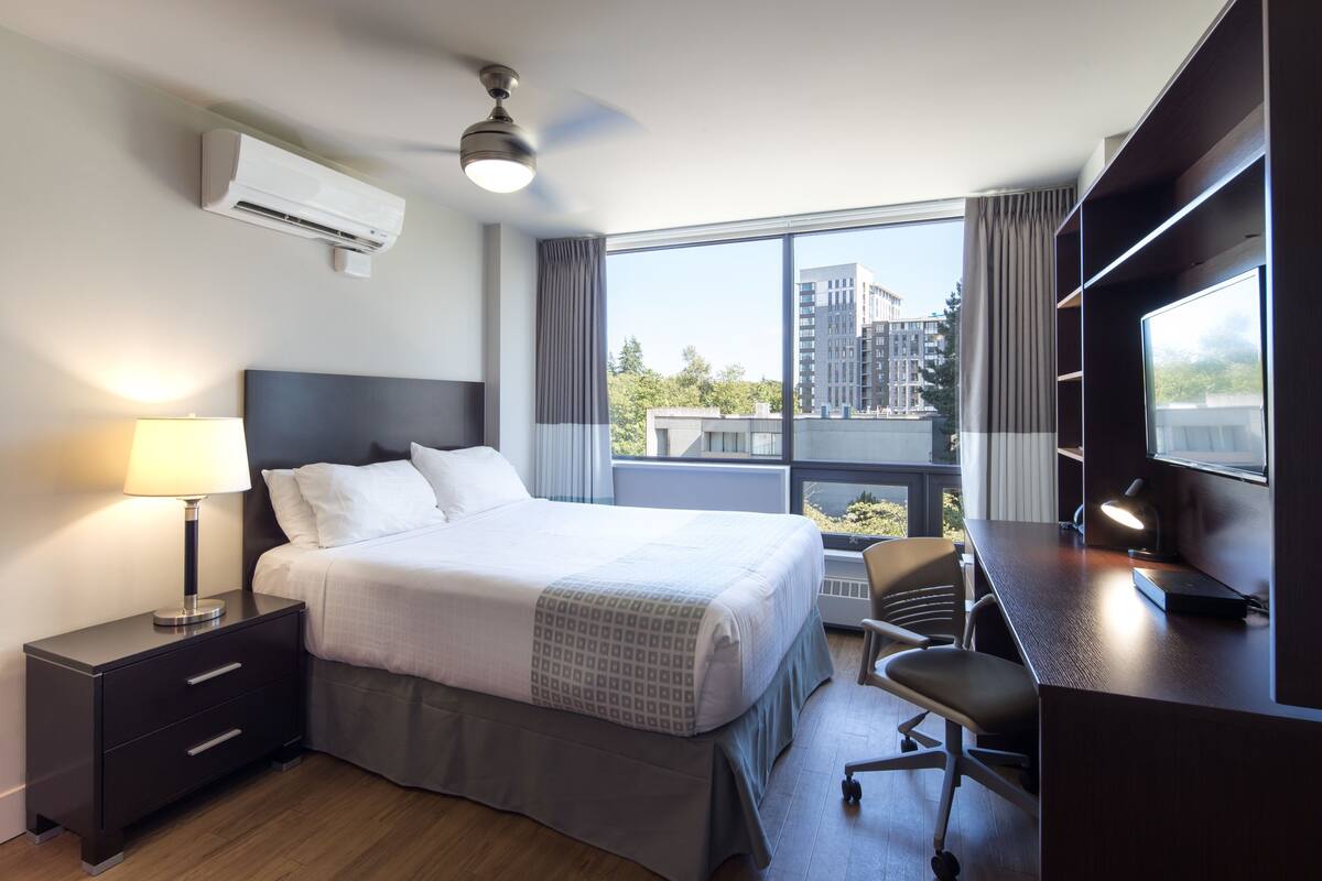 A hotel room with a bed, bedside table, lamp, desk with chair, and a large window with a view of buildings.