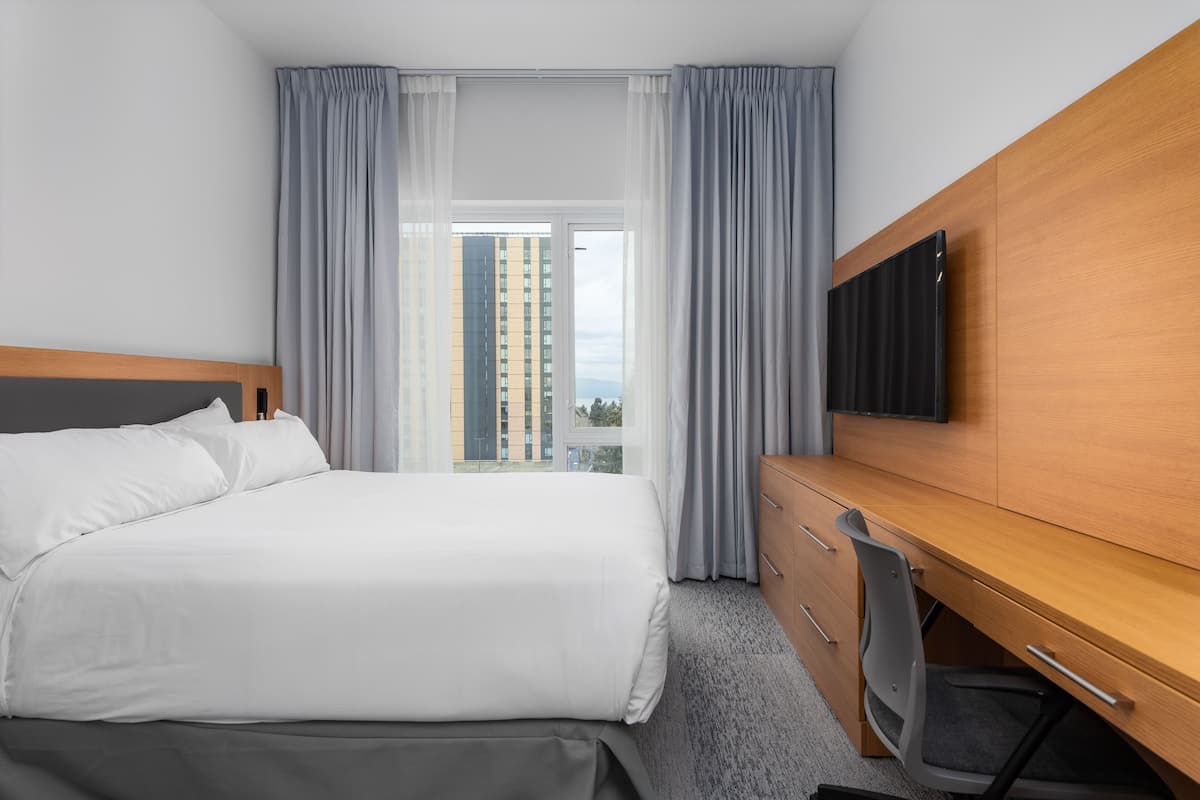A hotel room with a bed, a desk, a chair, a TV, and a view of a tall building.