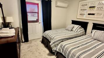 Une chambre avec deux lits, une commode, un miroir, une fenêtre avec des stores et un appareil de climatisation.