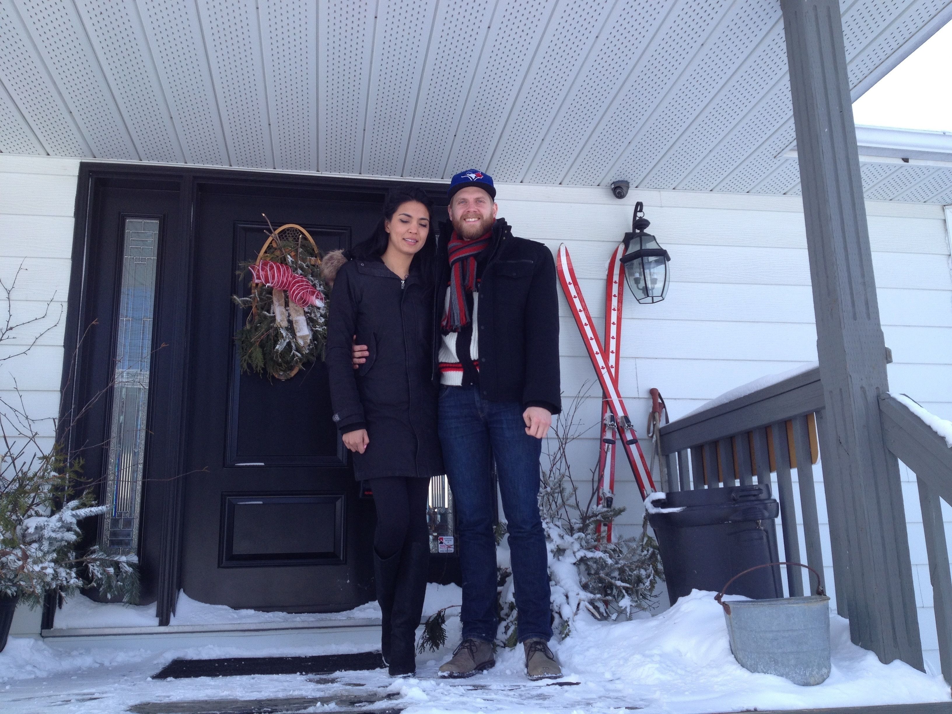 A couple standing in front of a house with a black door, a wreath, and a red and white ski pole leaning against the wall.