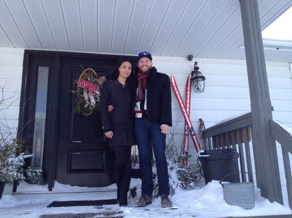 A couple standing in front of a house with a black door, a wreath, and a red and white ski pole leaning against the wall.