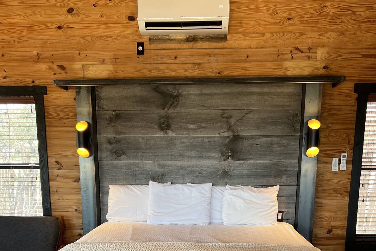 Ein Bett mit weißer, strukturierter Bettdecke, einem hölzernen Kopfteil mit integrierter Beleuchtung und einer wandmontierten Klimaanlage.