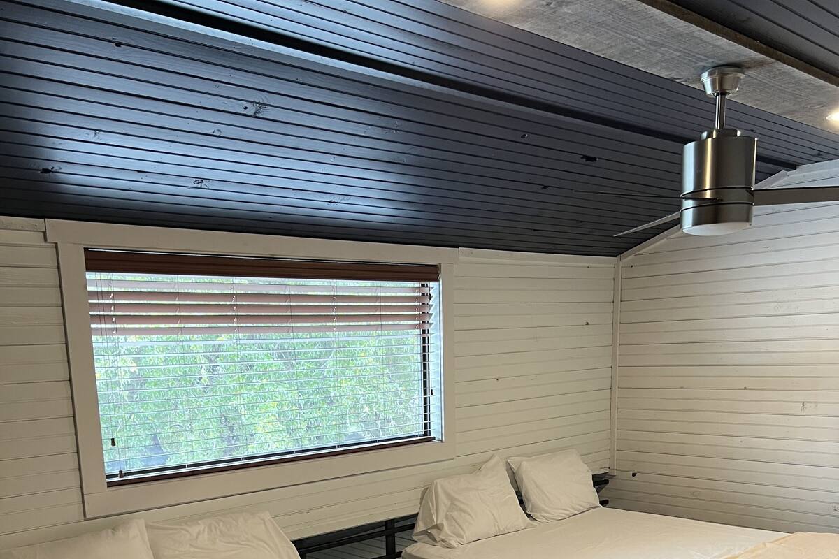Ein Zimmer mit zwei Betten, einem Fenster mit Jalousien, einem Deckenventilator und Holzböden.