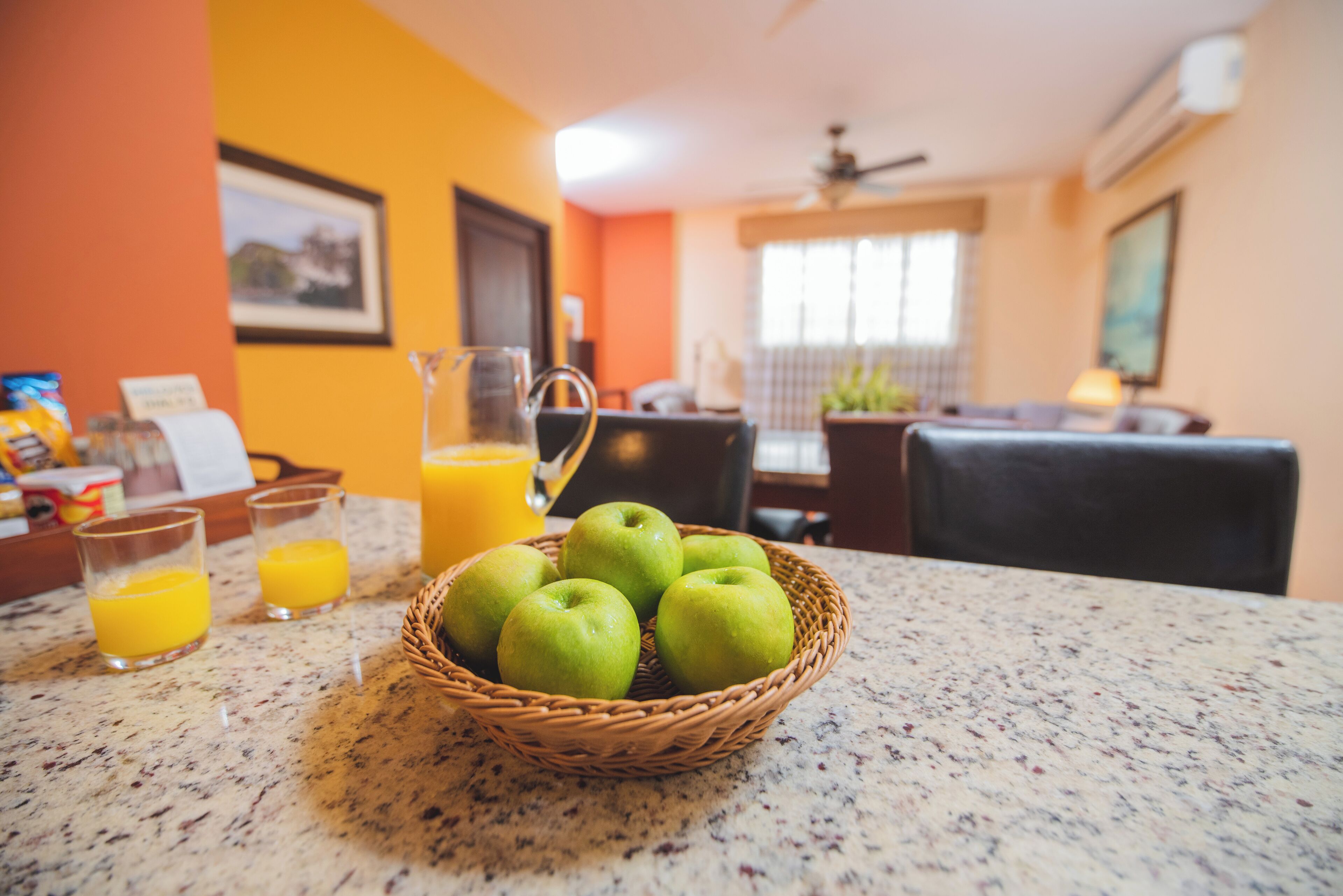 A kitchen counter with a basket of green apples, two glasses of orange juice, and a pitcher of water.