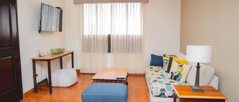 Una sala de estar con un sofá, una mesa de centro, una mesita auxiliar, un ventilador de techo, una ventana con cortinas y un televisor montado en la pared.