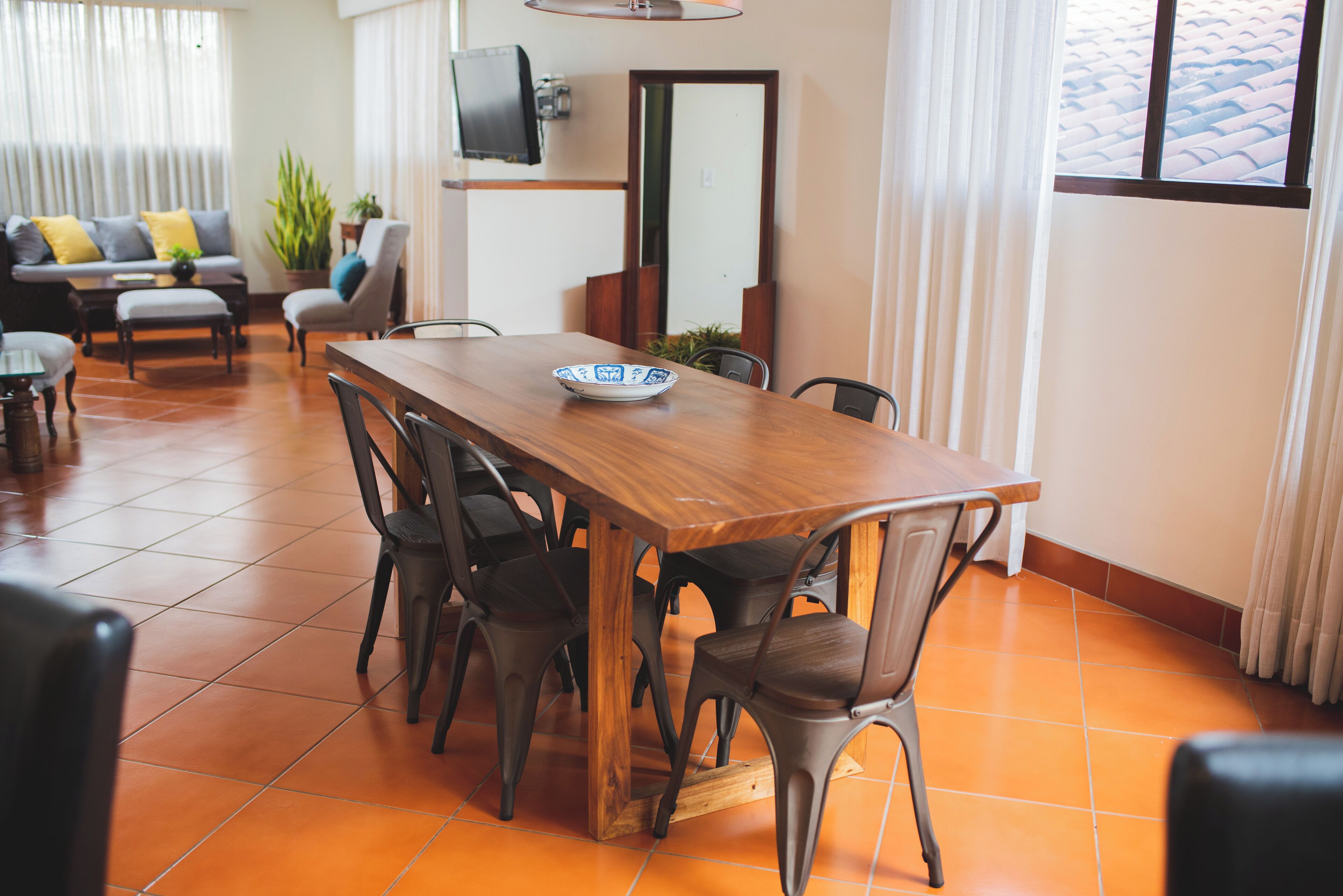 Un comedor con una mesa y sillas de madera, un televisor montado en la pared y un sofá al fondo.