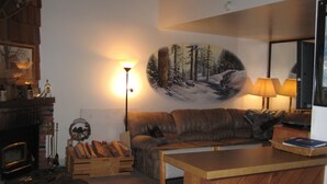 A cozy living room with a fireplace, a wooden table, a couch, and a painting of a forest scene on the wall.